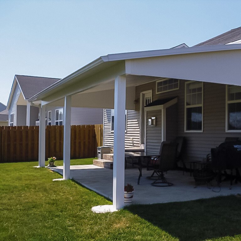 White open-sided patio cover with support posts and a solid roof over a backyard seating area.